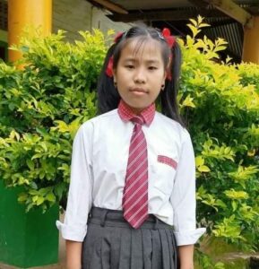 A girl in school uniform standing next to bushes.