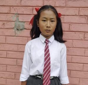 A young girl in school uniform standing against a brick wall.