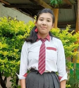 A girl in school uniform standing next to bushes.