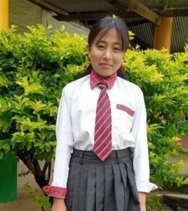 A girl in school uniform standing next to bushes.