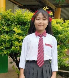 A girl in school uniform standing next to bushes.