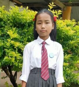 A girl in school uniform standing next to a bush.