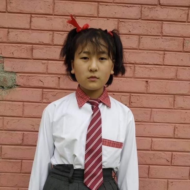 A young girl in school uniform standing against a brick wall.